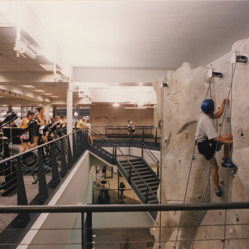 Multiple-story view of a climber on a rock wall to the right and people using exercise equipment on the balcony area to the left