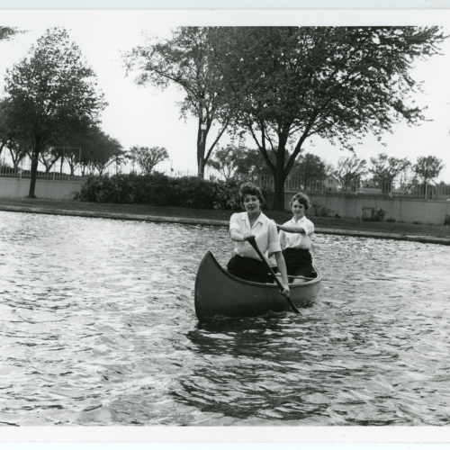 Two women wearing physical education uniforms canoeing on Prexy's Pond