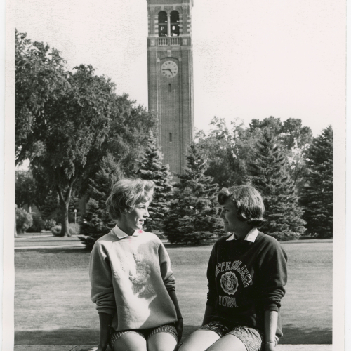 Two women seated, talking, on a brick wall with Campanile in background