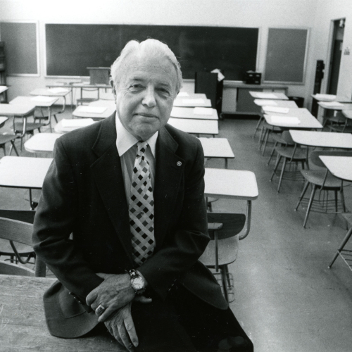 Man in jacket and tie sitting on desk at back of a classroom full of empty student desks