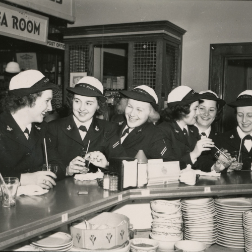 Six women in Navy uniforms havaing drinks at a counter; a sign partially visible behind them reads "Tea Room" and "Post Office"