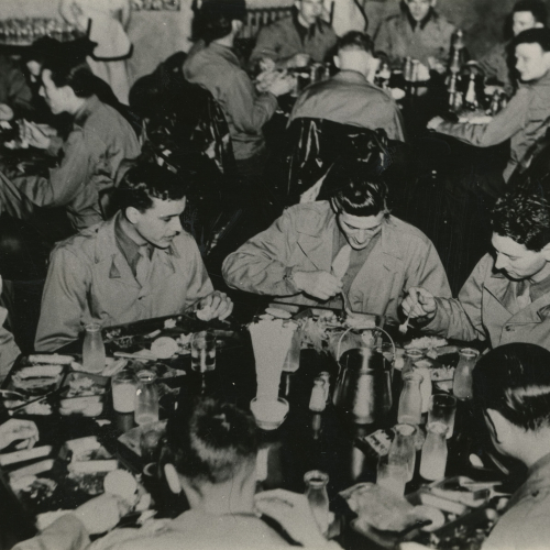 Men of the Army Air Corps in uniform, seated around three visible round tables eating