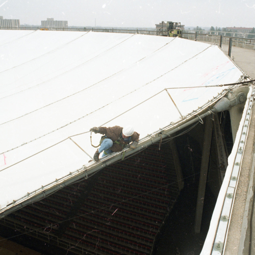 One man on rim of building where it meets deflated fabric roof, two other men watching from walkway