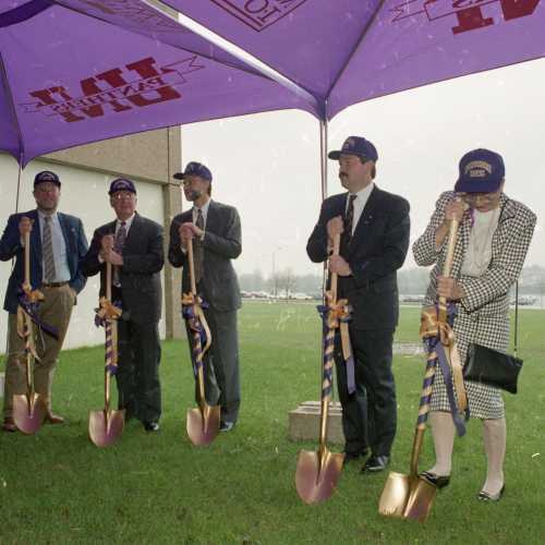 8 people in formal clothes, each wearing a hat and breaking ground with a purple-and-gold-ribbon-wrapped golden shovel outside under a tent
