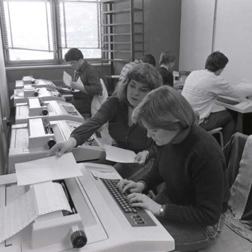 Two students working together in room with multiple typewriters, four other students working behind them