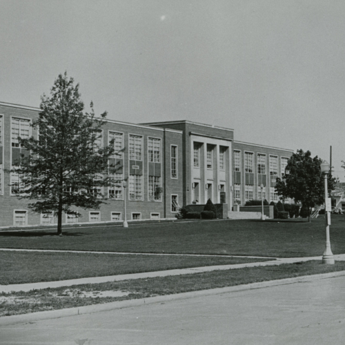 Two-story brick building with high windows, flat roof, and columns at the entrance