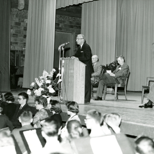 Man in suit speaking at podium on stage, three men seated behind, and audience and school band members in front of stage