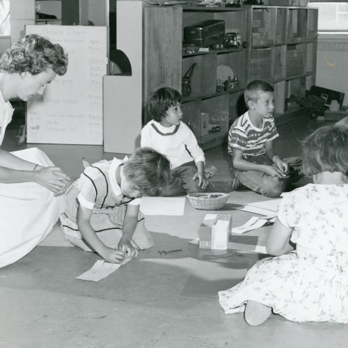 A woman crouched on the floor next to a circle of five children working on a paper craft project in a classroom