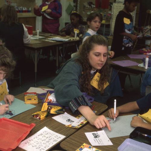 Teacher or student teacher helping two children with a craft project at a cluster of desks in a classroom; two other clusters of students in background