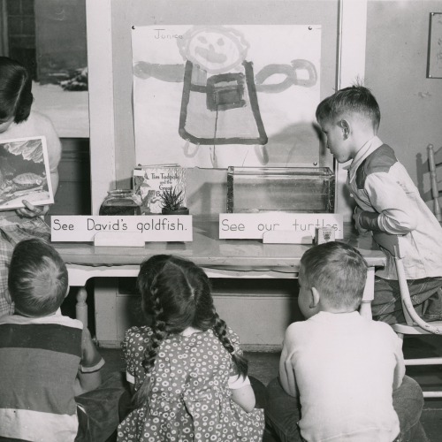 Woman reading picture book to four children, there are two tanks on a table in front of them, one labeled See David's goldfish and the other with See our turtles
