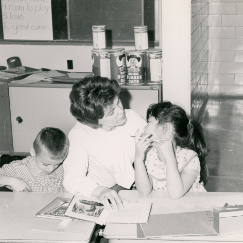Woman reading a book to a young girl and boy at a table; behind them is a counter with books and Quaker oats containers