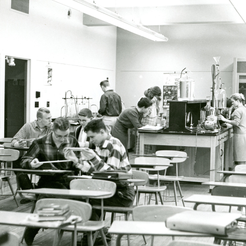 High school students in a science classroom, four studying at desks and about six running experiments at the workbenches in the back