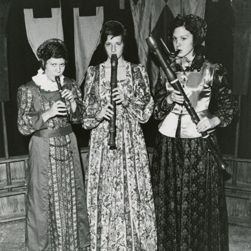 Three young women in medieval-style dresses in front of two tapestries playing versions of the record; left to right, small recorder, larger clarinet-size recorder, and bassoon-size recorder
