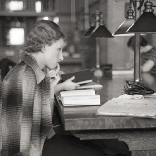 Woman studying at library table, book and notebooks in front of her and finger at her lips
