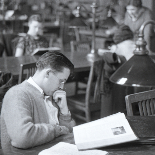 Man reading a book at library table, other students reading at tables in background
