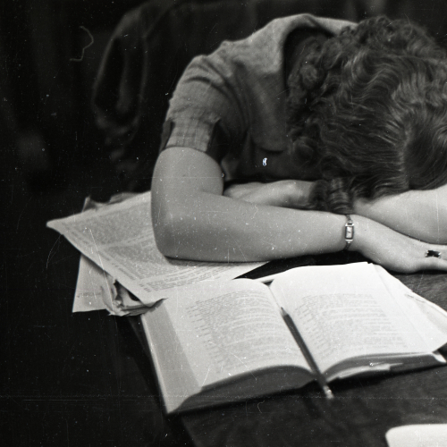 Woman sleeping with head down on her arms crossed on the table in front of her, books and notes scattered