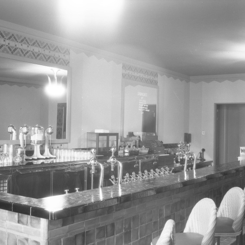 Empty soda fountain counter with taps, register, glasses and other miscellany on one side and bar chairs on the other