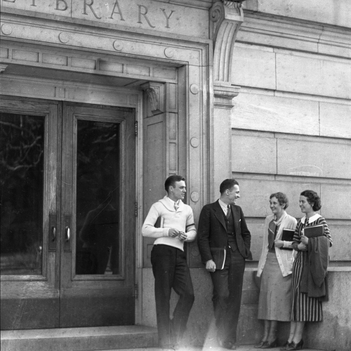 Two men and two women talking to each other on the steps of a building with the word Library on the lintel