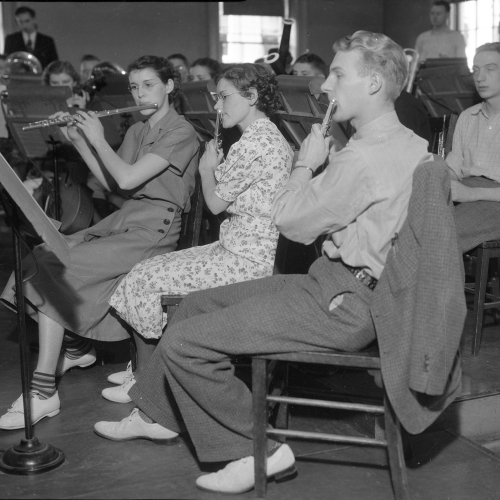 Room of seated students playing musical instruments, focus on four flute players in first row
