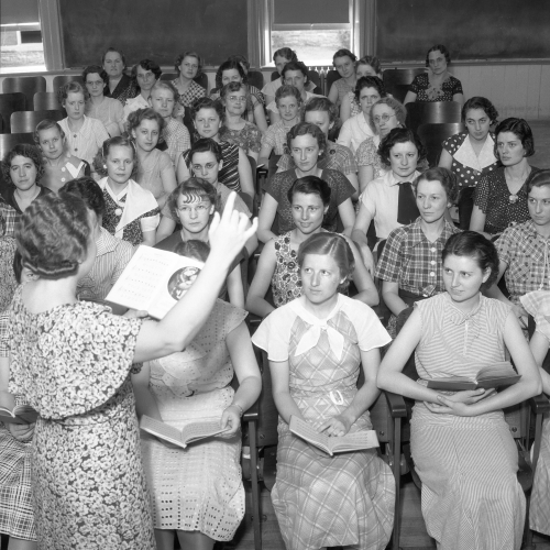 Professor directing group of approximately forty seated women who hold choral books